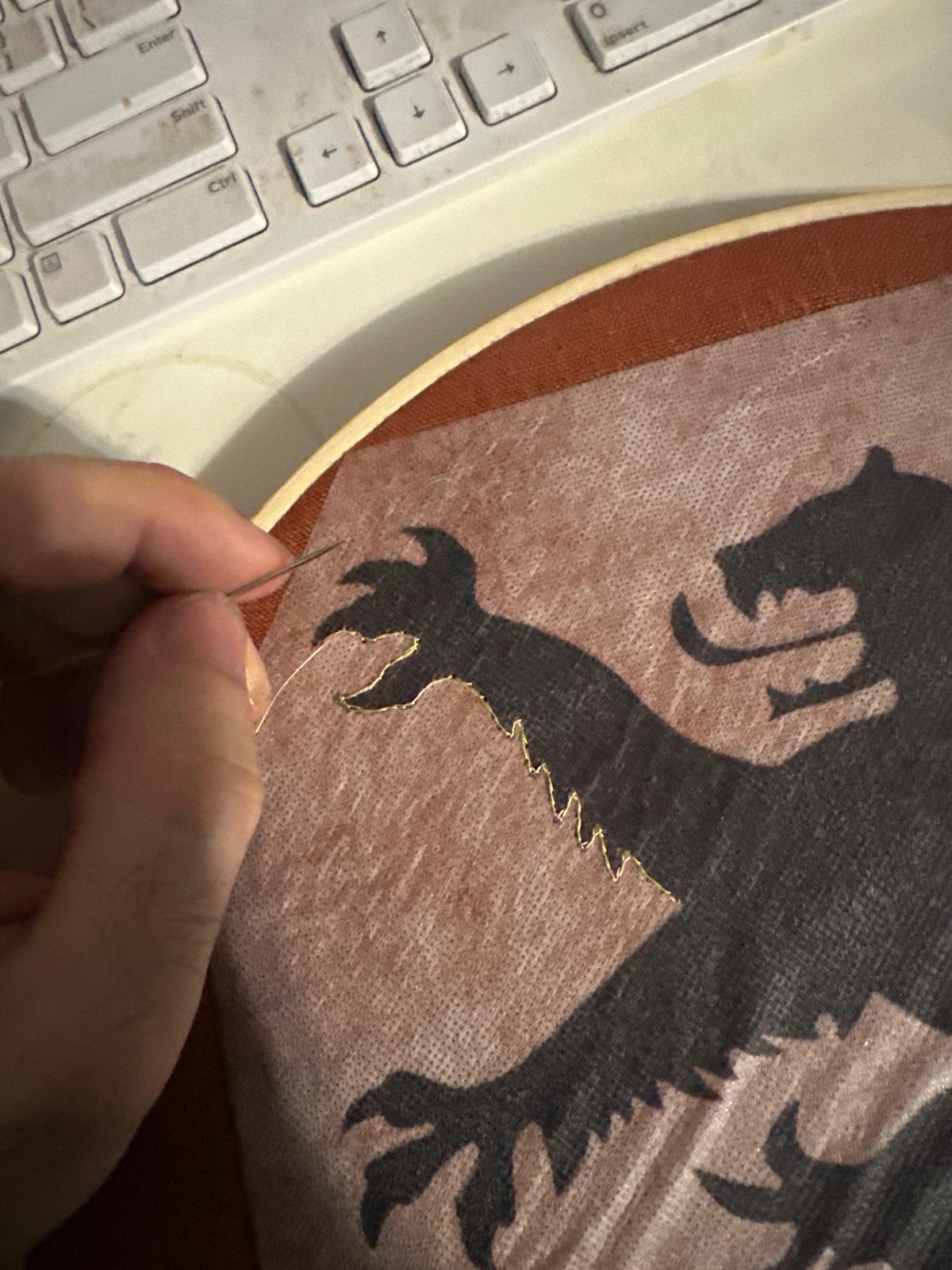 A lion being embroidered on reddish fabric with gold thread